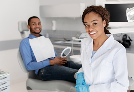 A dental hygienist wearing a white coat and blue gloves is smiling at a patient seated in a chair with a dental mirror and light, while a man in a suit looks on.